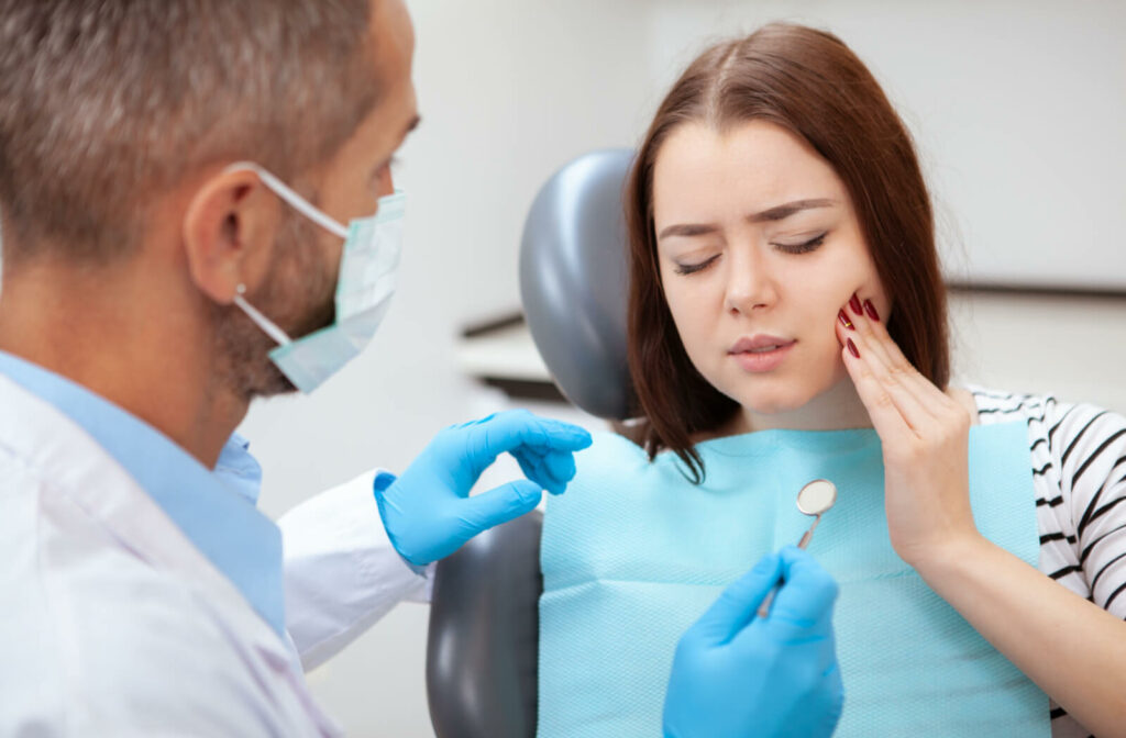 A woman with a toothache is sitting in a dental chair, touching her cheek area where the aching tooth is. While a male dentist is preparing to examine with a dental tool.