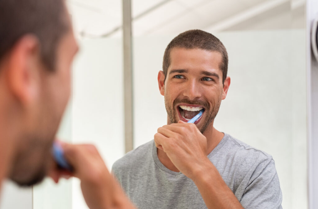 A happy young man in a grey shirt brushing his teeth in front of a mirror.